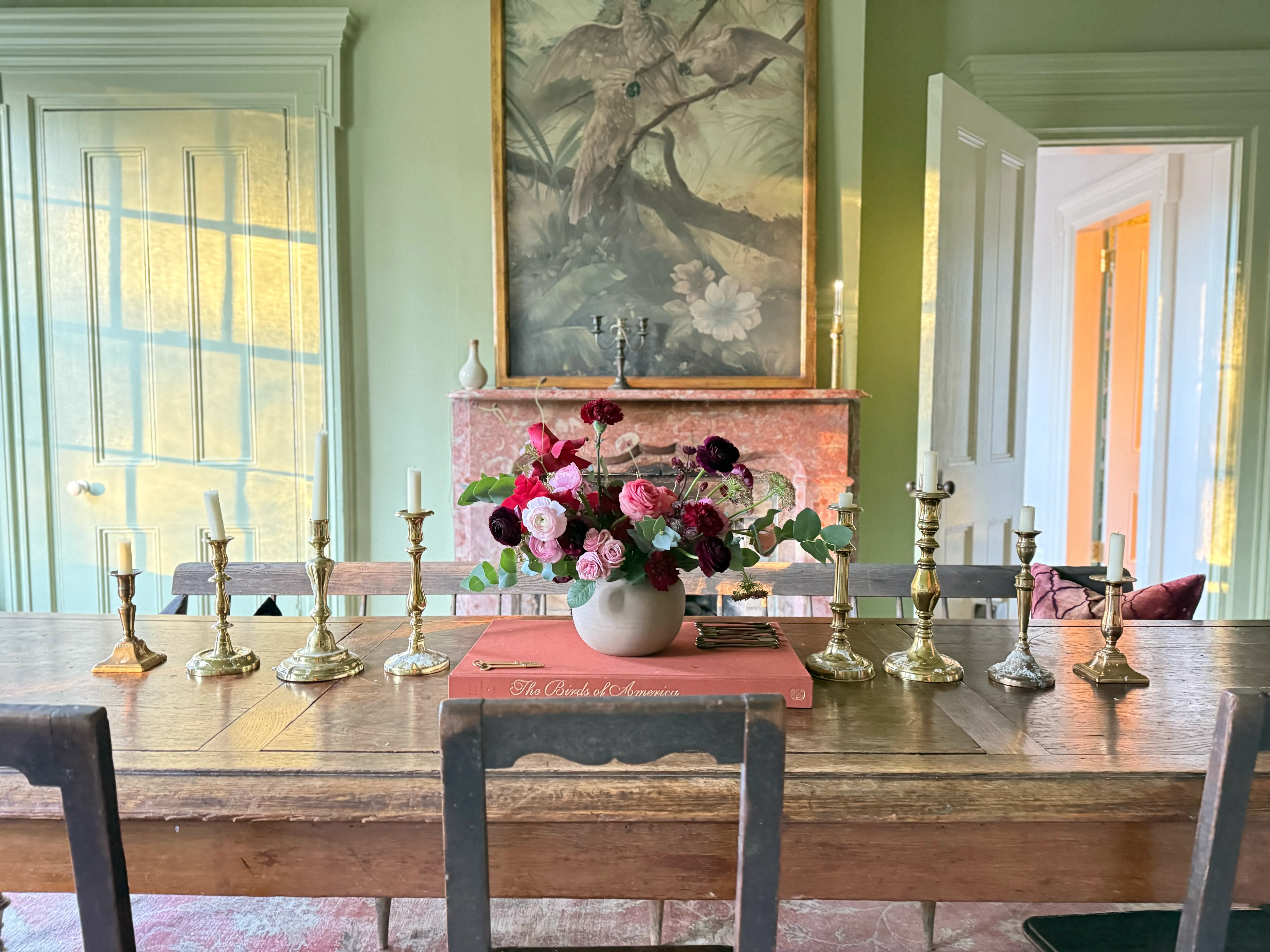 Set dining table in a sage-green room with floral centerpiece, brass candlesticks, and late afternoon light.